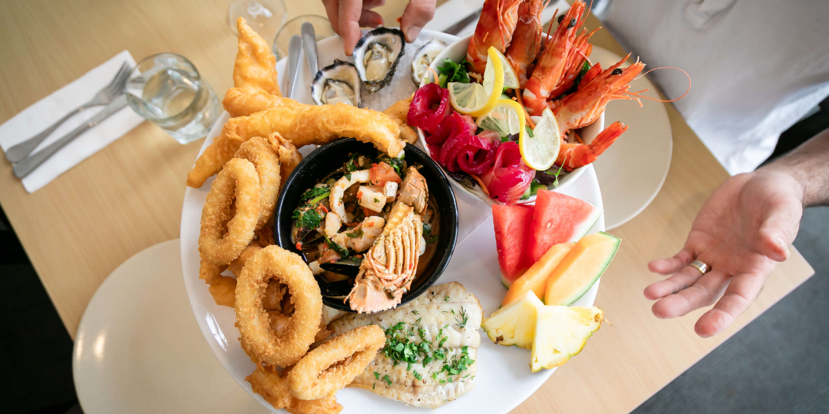 Assorted seafood and vegetable, fruits platter on a table with hands reaching towards it.