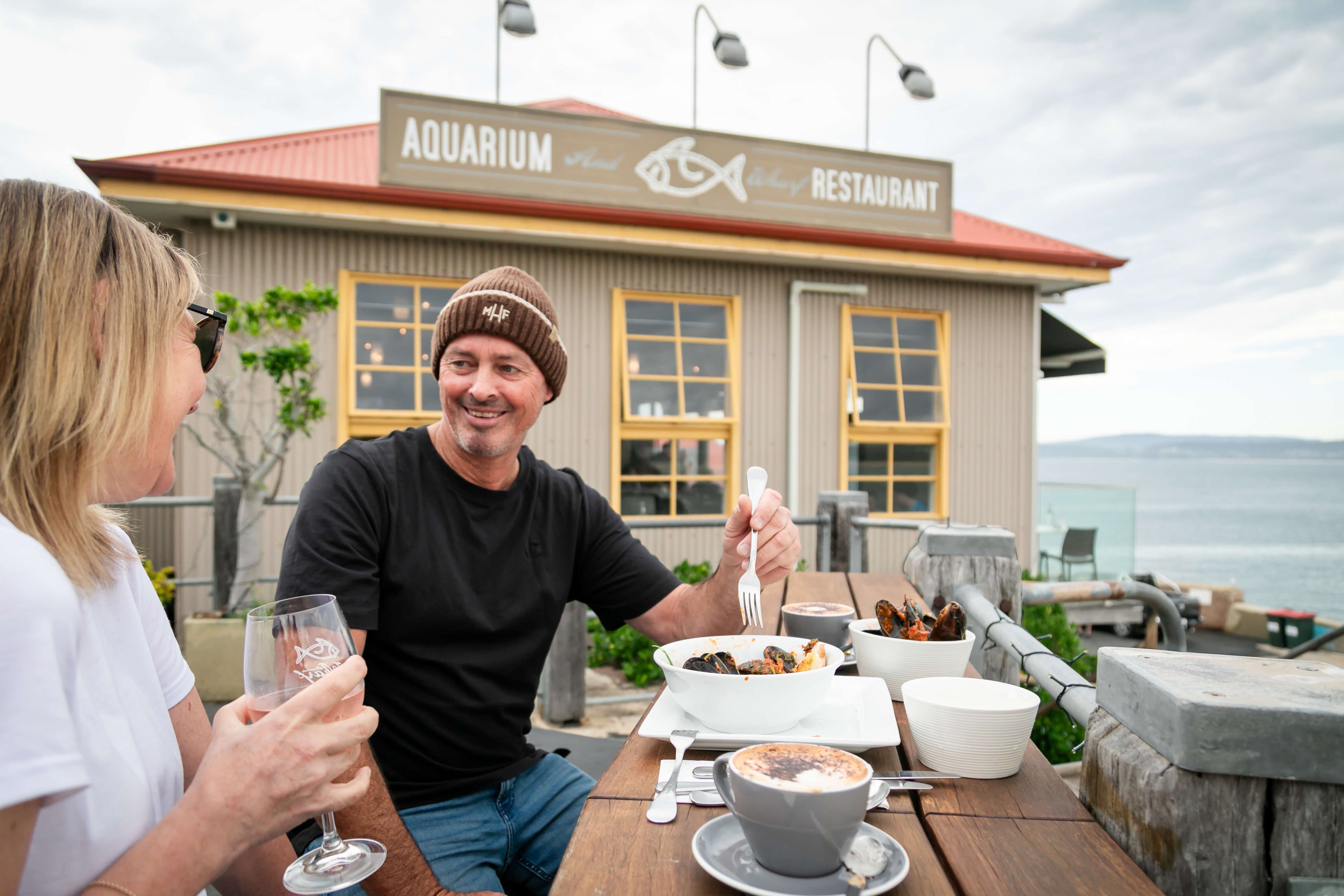 Two people enjoying a meal outdoors at an aquarium restaurant with a scenic background. Merimbula Restaurant