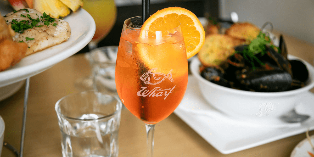 Glass of orange cocktail with Merimbula Wharf branding on a table with food.
