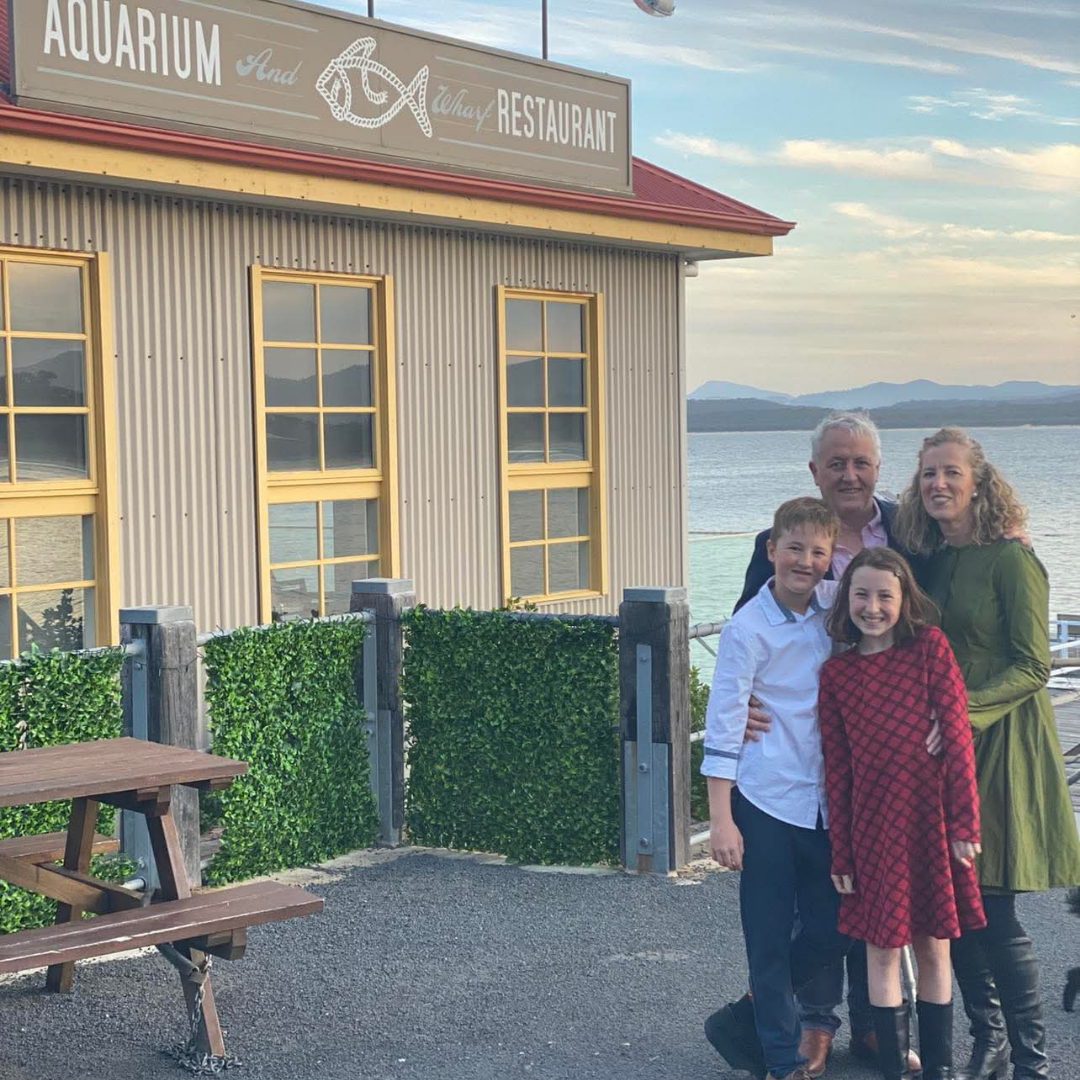 Merimbula Wharf Family of four standing in front of a restaurant named 'Aquarium and Wharf Restaurant' by the Merimbula waterfront. Sapphire Coast