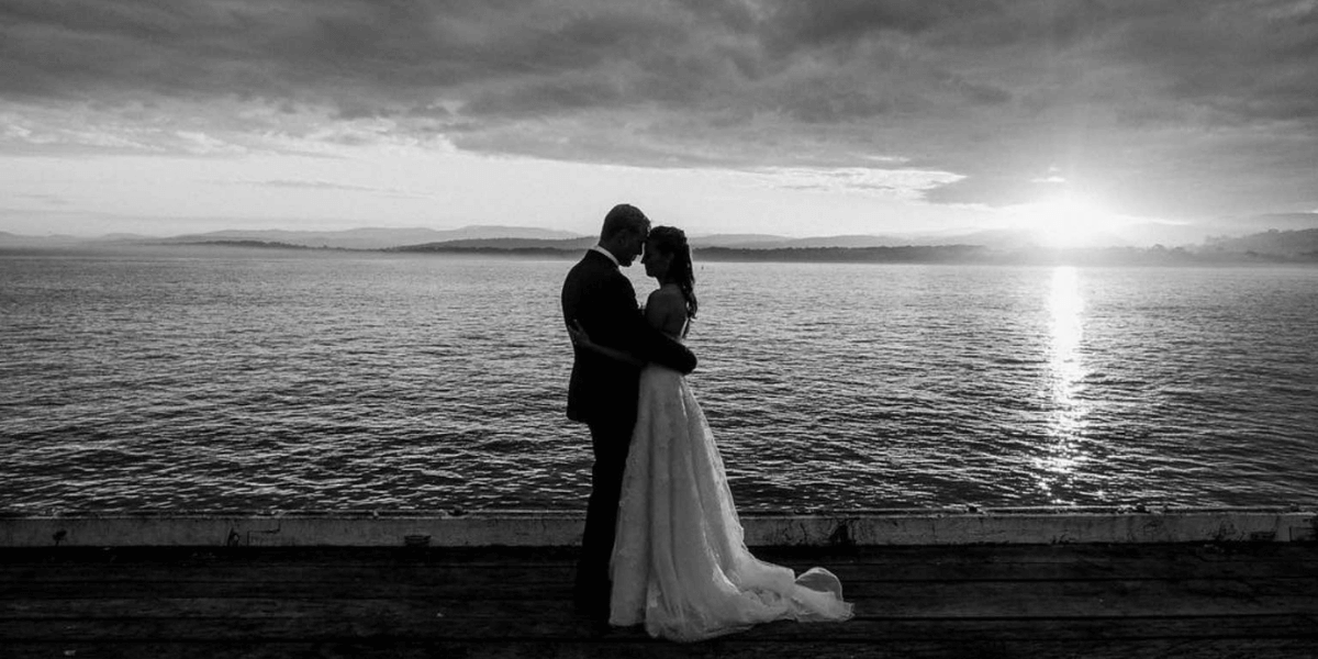 Black and white image of a couple embracing by a body of water with a sunset.