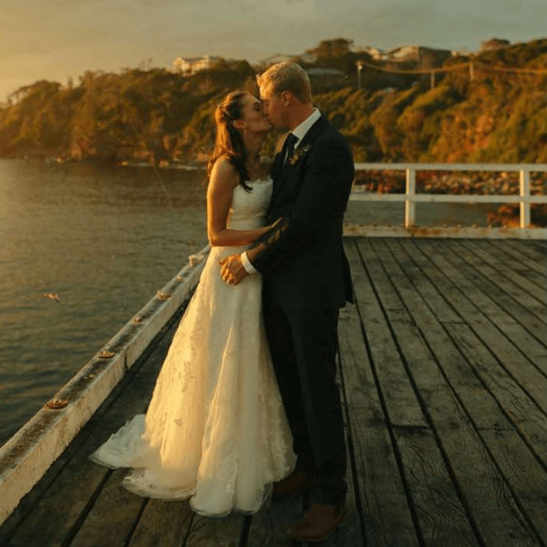 Wedding couple kissing on a wooden pier by a body of water at sunset.