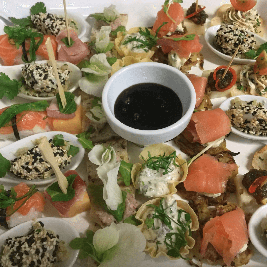 Assorted canapés with salmon and vegetables on a white plate with a small bowl of sauce.