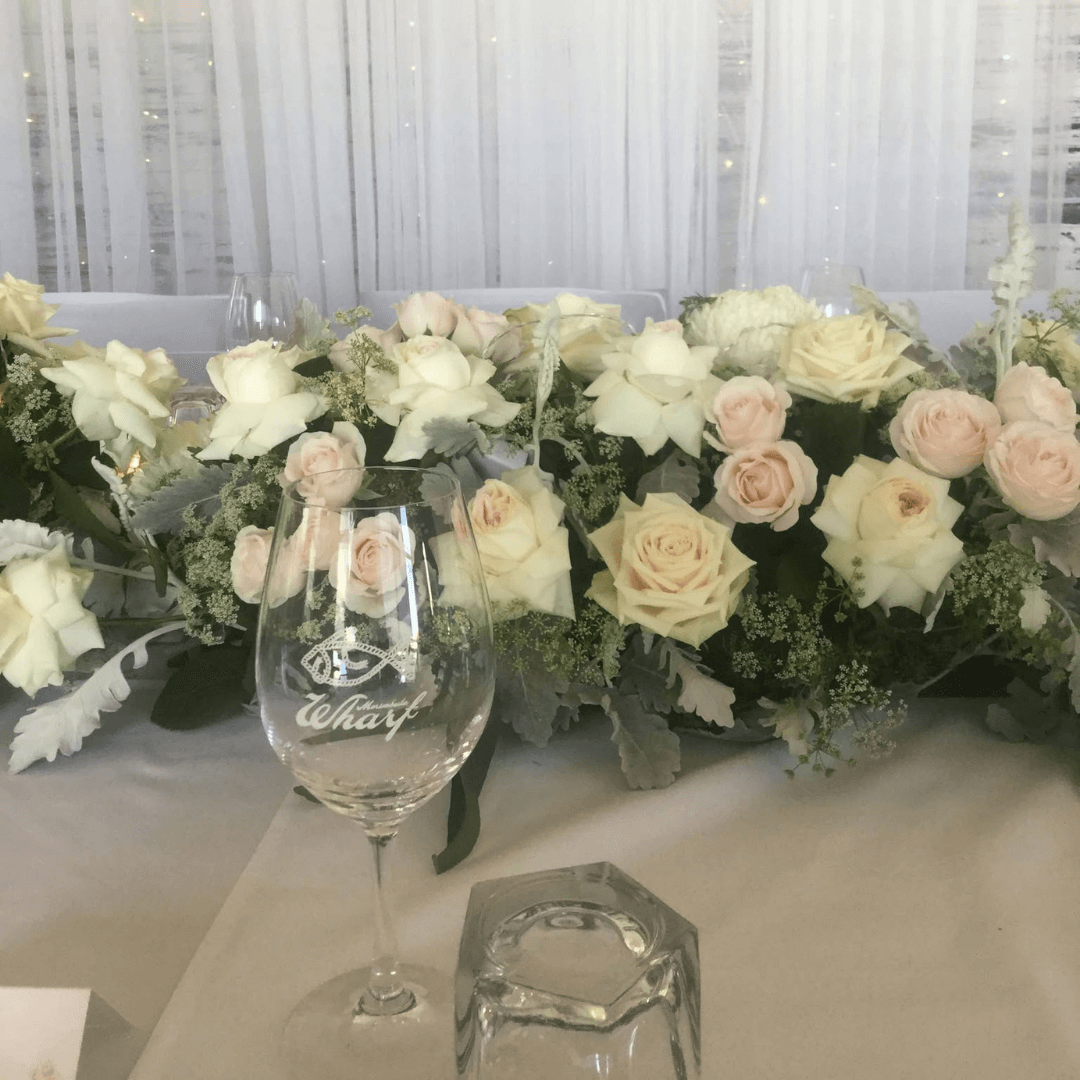 Decorative floral arrangement with a glass containing the Merimbula Warf Restaurant logo on a table, adorned for a wedding.