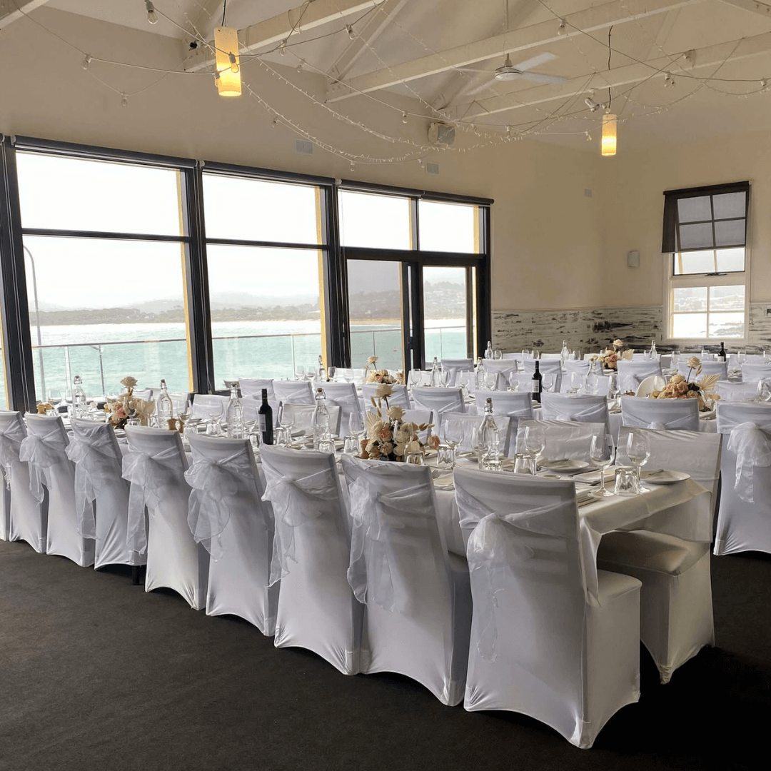 Long table set for a formal event with white tablecloths and chairs in a room with large windows.