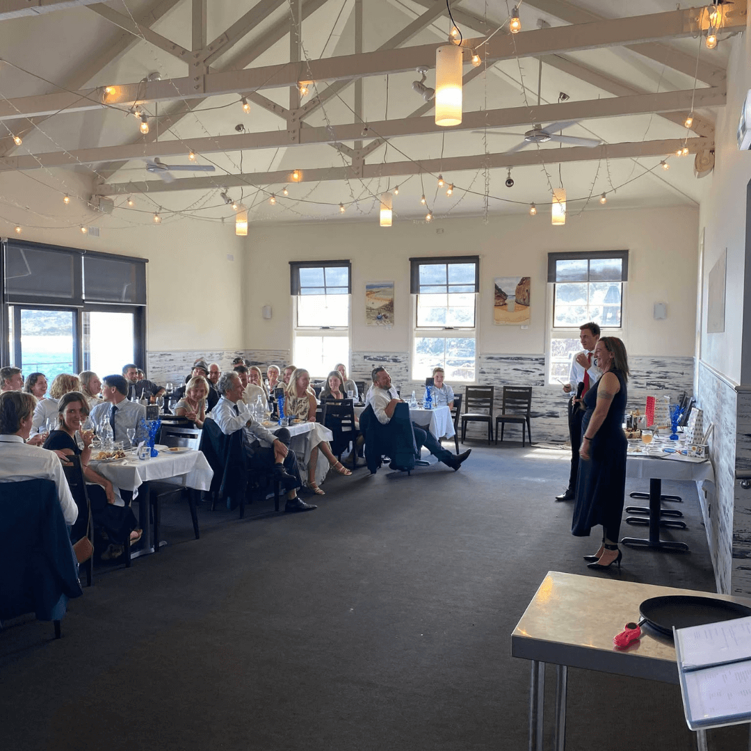 Group of people attending wedding, listening to speeches, sitting at tables in a large room with high ceilings and large windows. Merimbula Aquarium and Wharf Restaurant