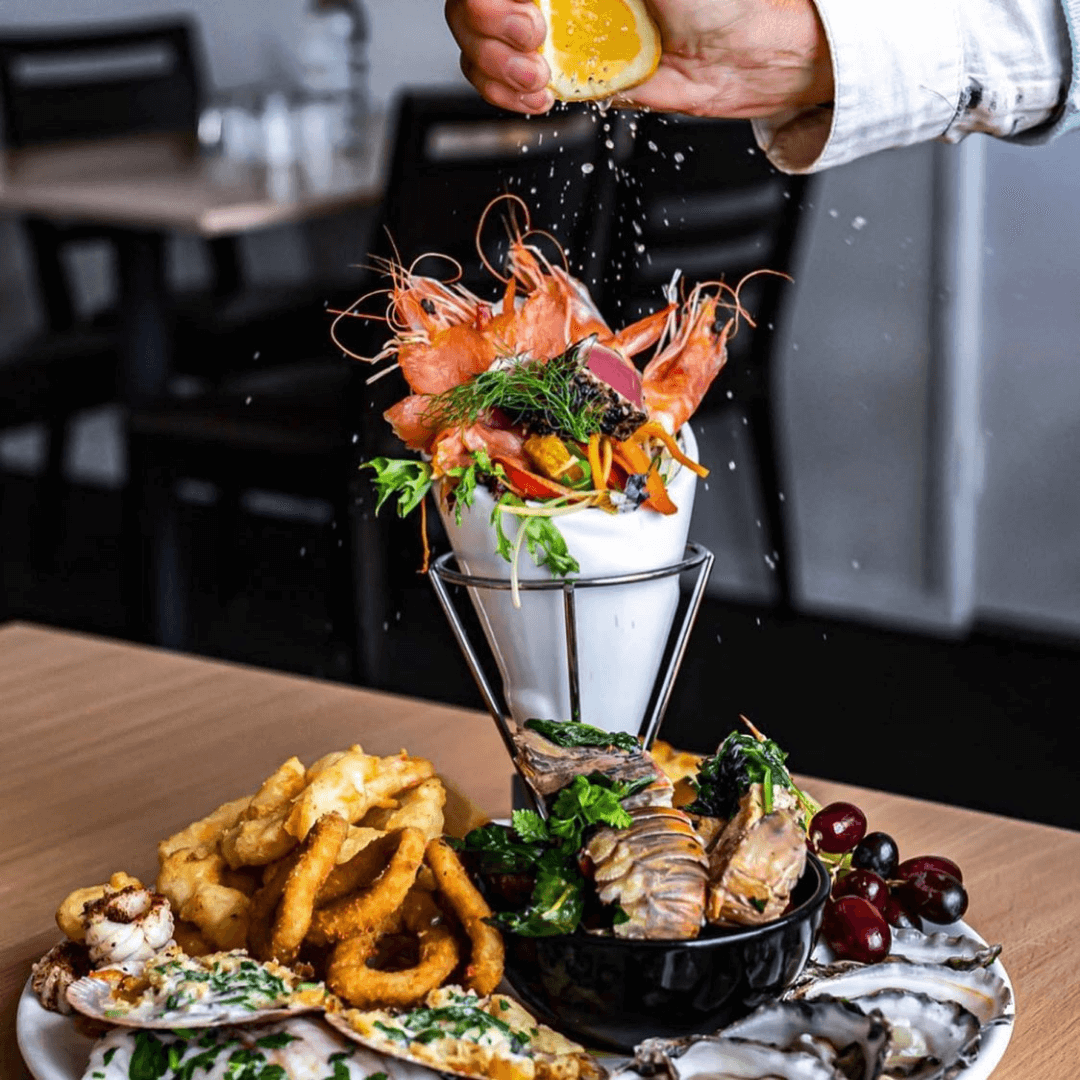 Seafood platter with a cone of seafood being garnished with a lemon wedge, set on a wooden table. Merimbula Restaurant