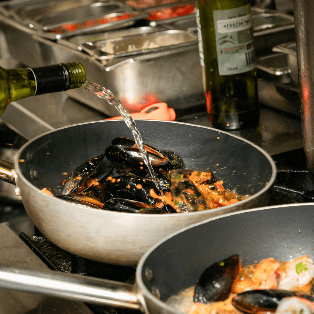 Mussels being cooked in a pan with wine being poured over them, surrounded by kitchen equipment. Merimbula Restuarant