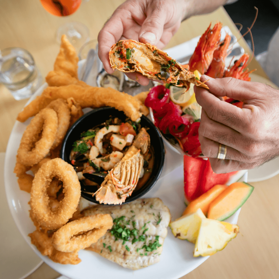 Assorted seafood and fried items on a plate with a person picking up food. Merimbula Restuarant