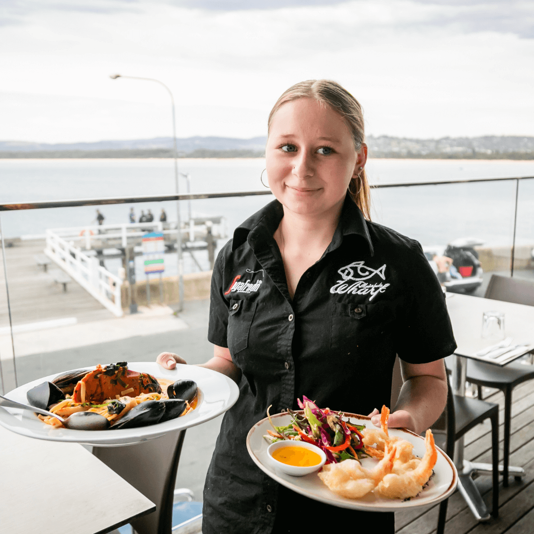 Person holding two plates of food on a balcony with a scenic view. Merimbula Restuarant