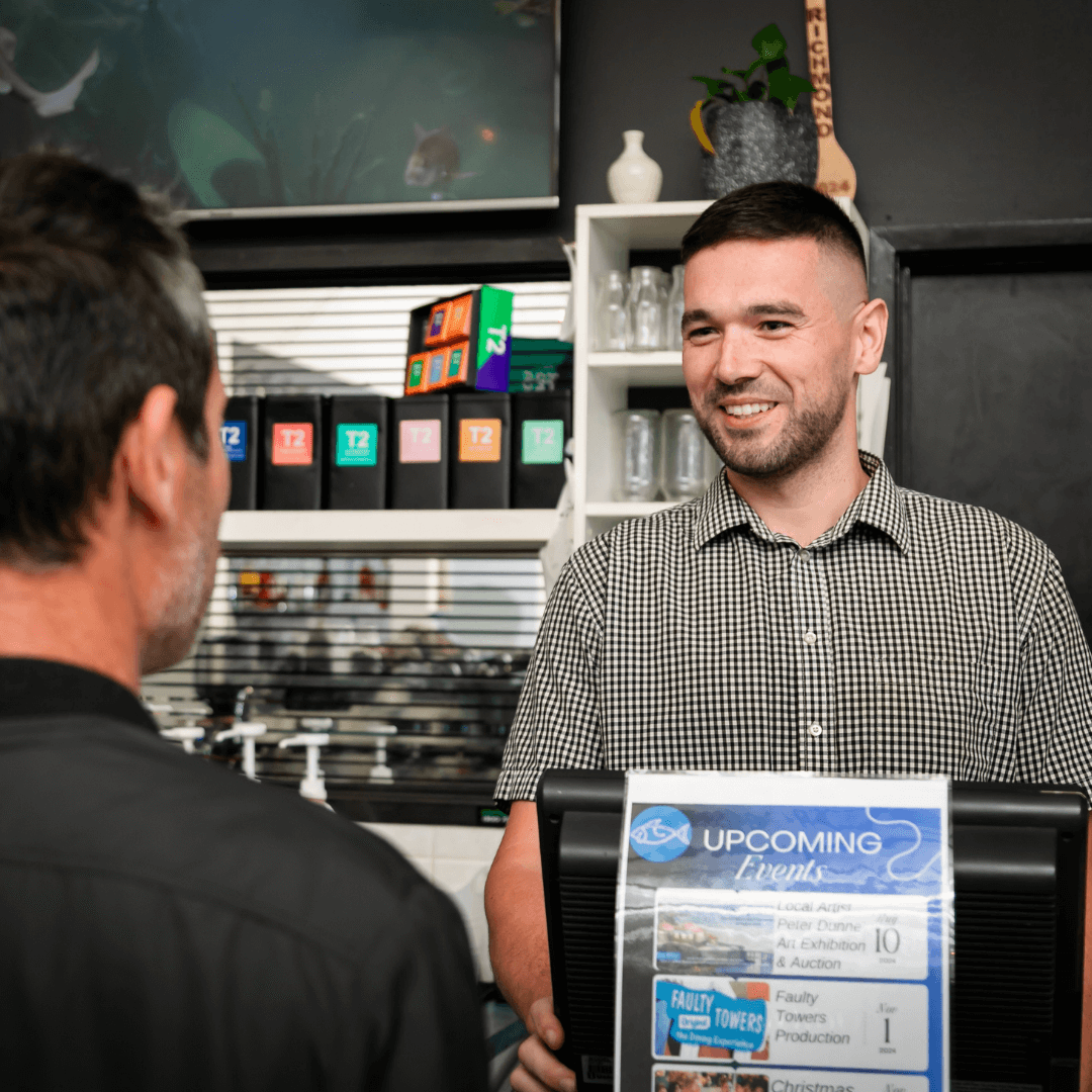 Man behind a counter with a customer, smiling, in a coffee shop setting. Merimbula  Restaurant