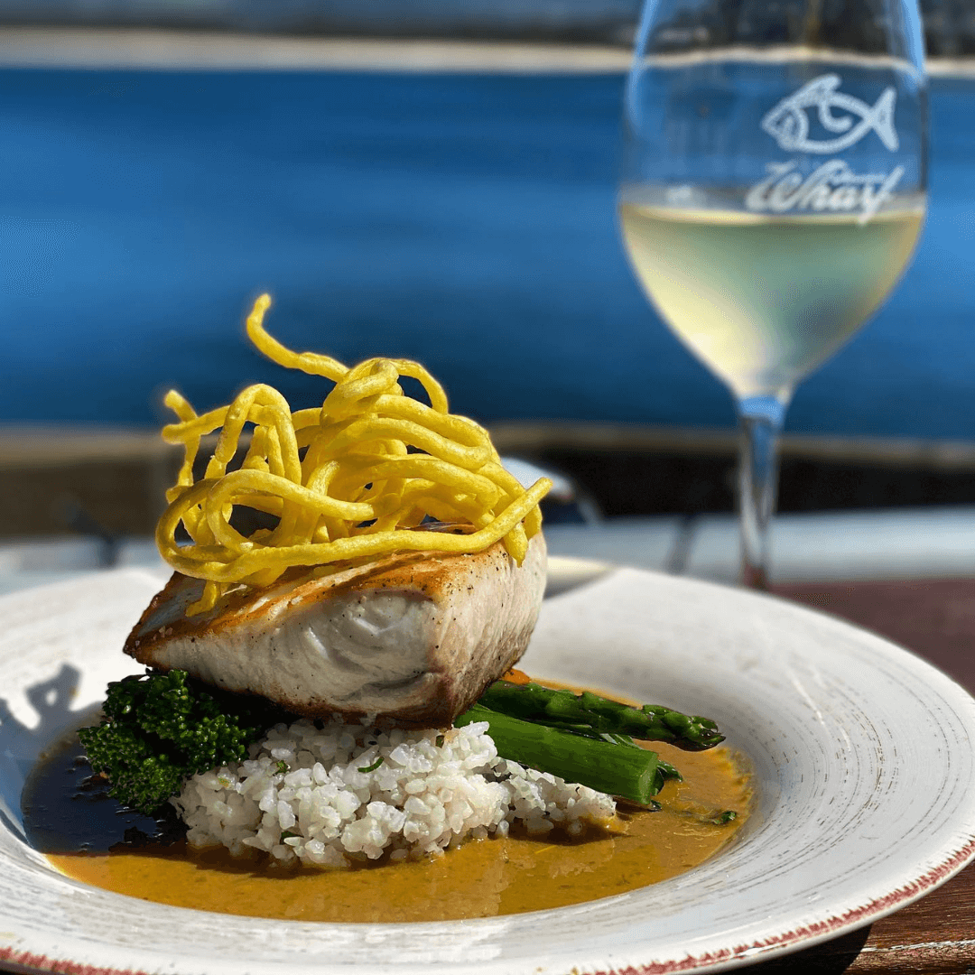 Plated dish with meat, vegetables, and noodles next to a glass of white wine on a blurred outdoor background. Merimbula Restaurant