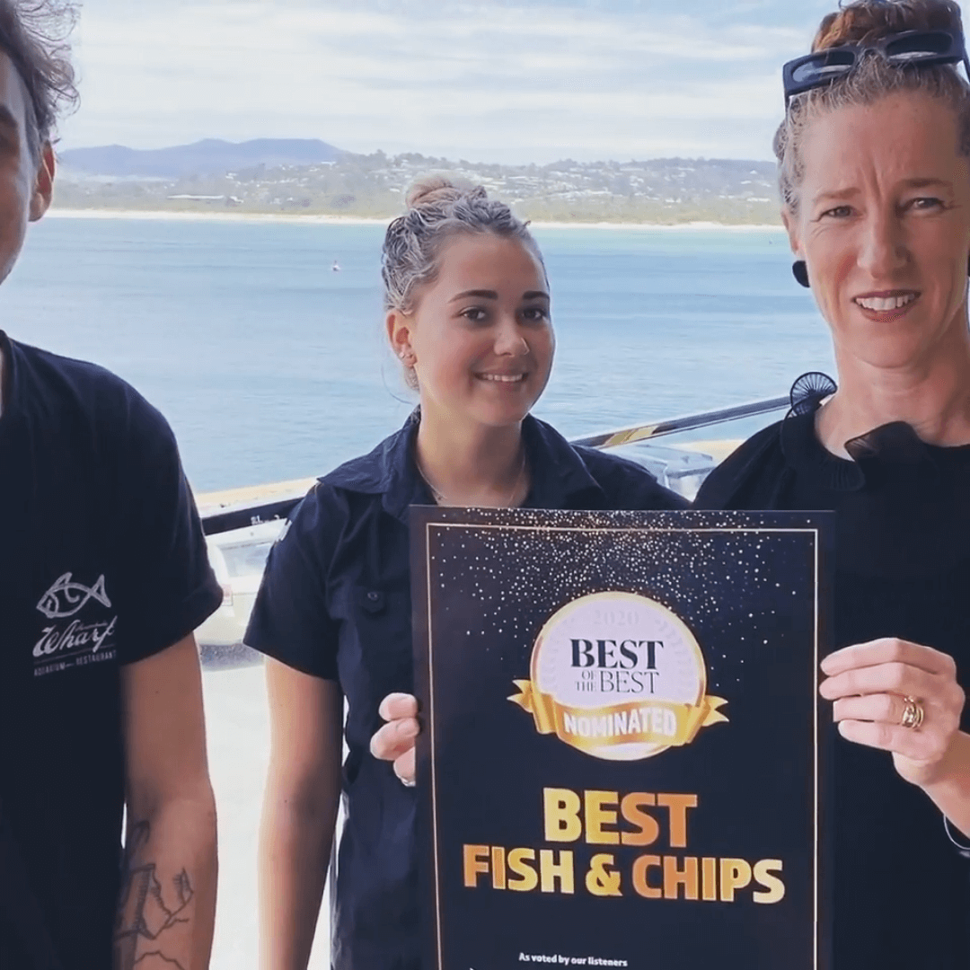 Three people holding a 'Best Fish & Chips' sign by a waterfront.