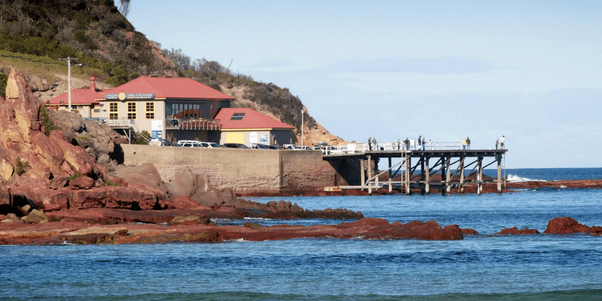 Coastal scene with a Merimbula Wharf Aquarium and Restaurant building, pier, and rocky shoreline.