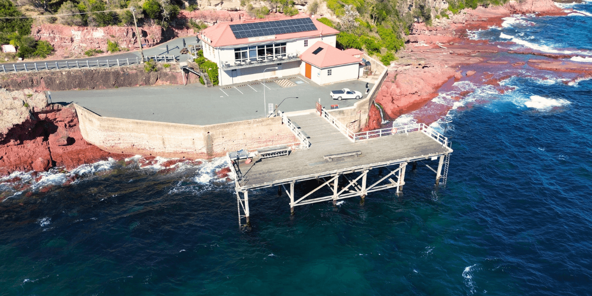 Aerial view of a pier extending into the ocean with rocky cliffs and buildings in the background.